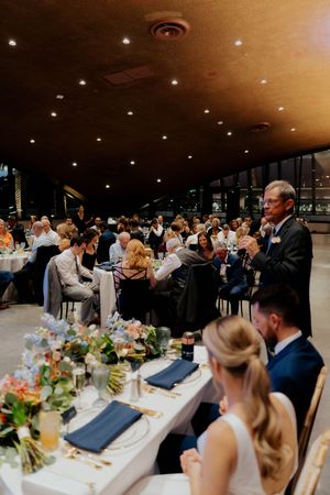 Wedding guests seated at elegant banquet tables with floral centerpieces enjoy dinner in a dimly lit reception hall.