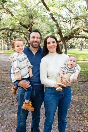 Smiling family stands together under oak trees in a park on an autumn day wearing casual denim and sweaters.