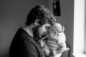 A tender black and white portrait of a bearded father in dark clothing holding an infant by a window.