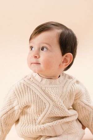 Small child wearing cozy cream-colored cable knit sweater gazes upward against soft pink background.