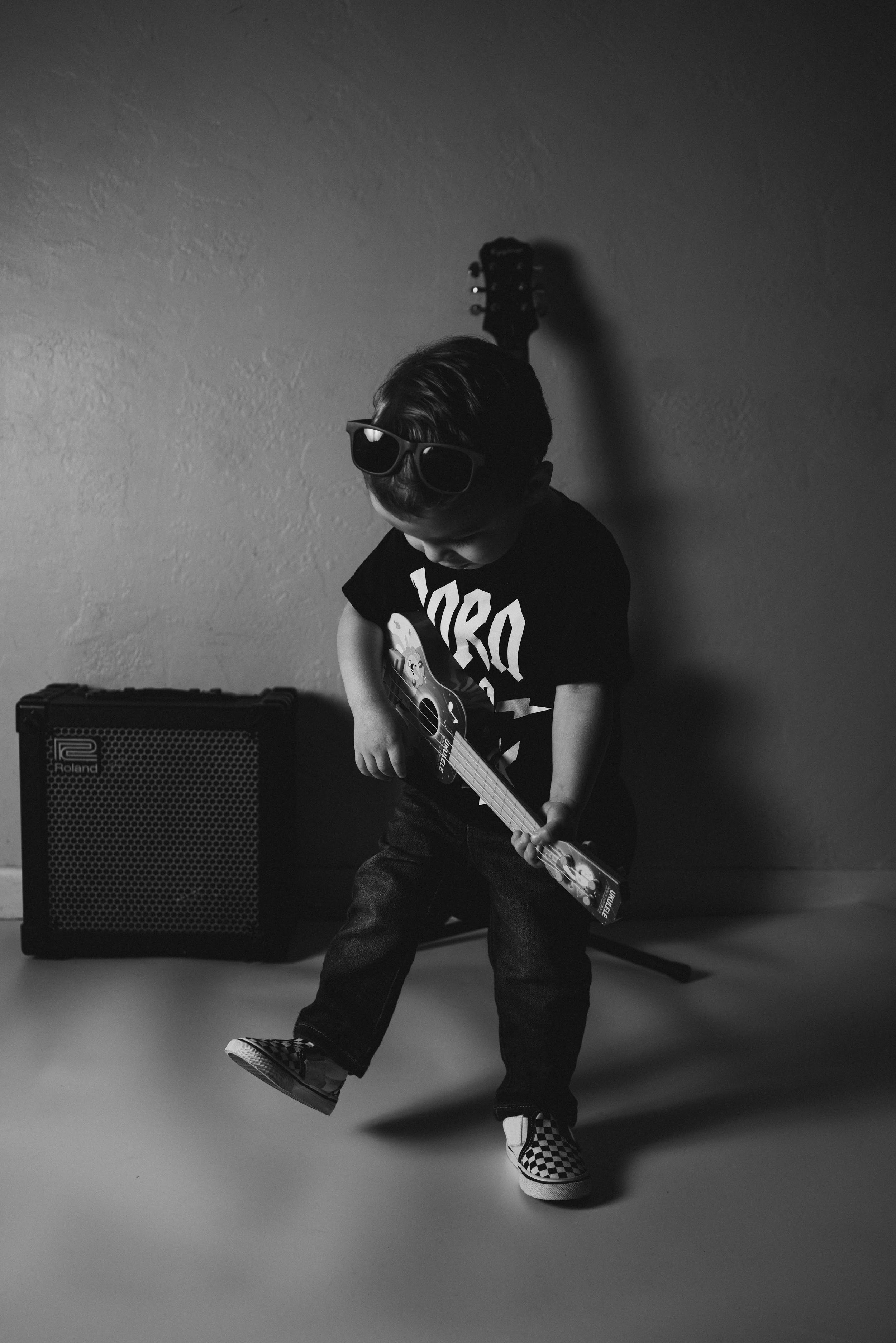 A young musician in a black t-shirt plays electric guitar next to an amp in a moody black and white photo.