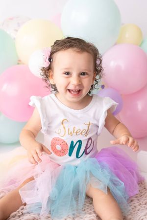 A baby wearing a 'Sweet One' shirt and rainbow tutu celebrates against a backdrop of pastel balloons.