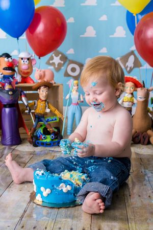 A baby in jeans enjoys a Toy Story themed cake smash photo session with balloons and character toys in the background.