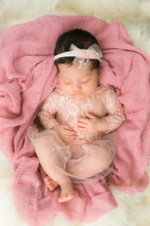 Sleeping baby wearing delicate lace dress and headband lying on pink textured blanket with white fur trim background.