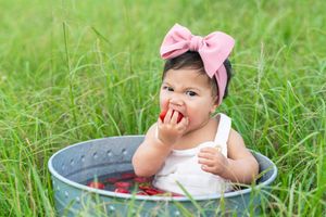 Small child wearing pink bow sits in grey tub filled with strawberries on grassy lawn.