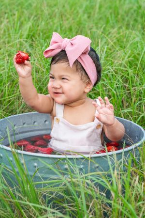 A small child wearing a pink bow sits in a metal tub filled with water and strawberries on grass.