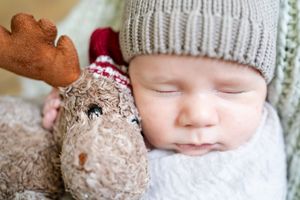 A sleeping baby bundled in white with a gray knit hat rests next to a plush reindeer toy.