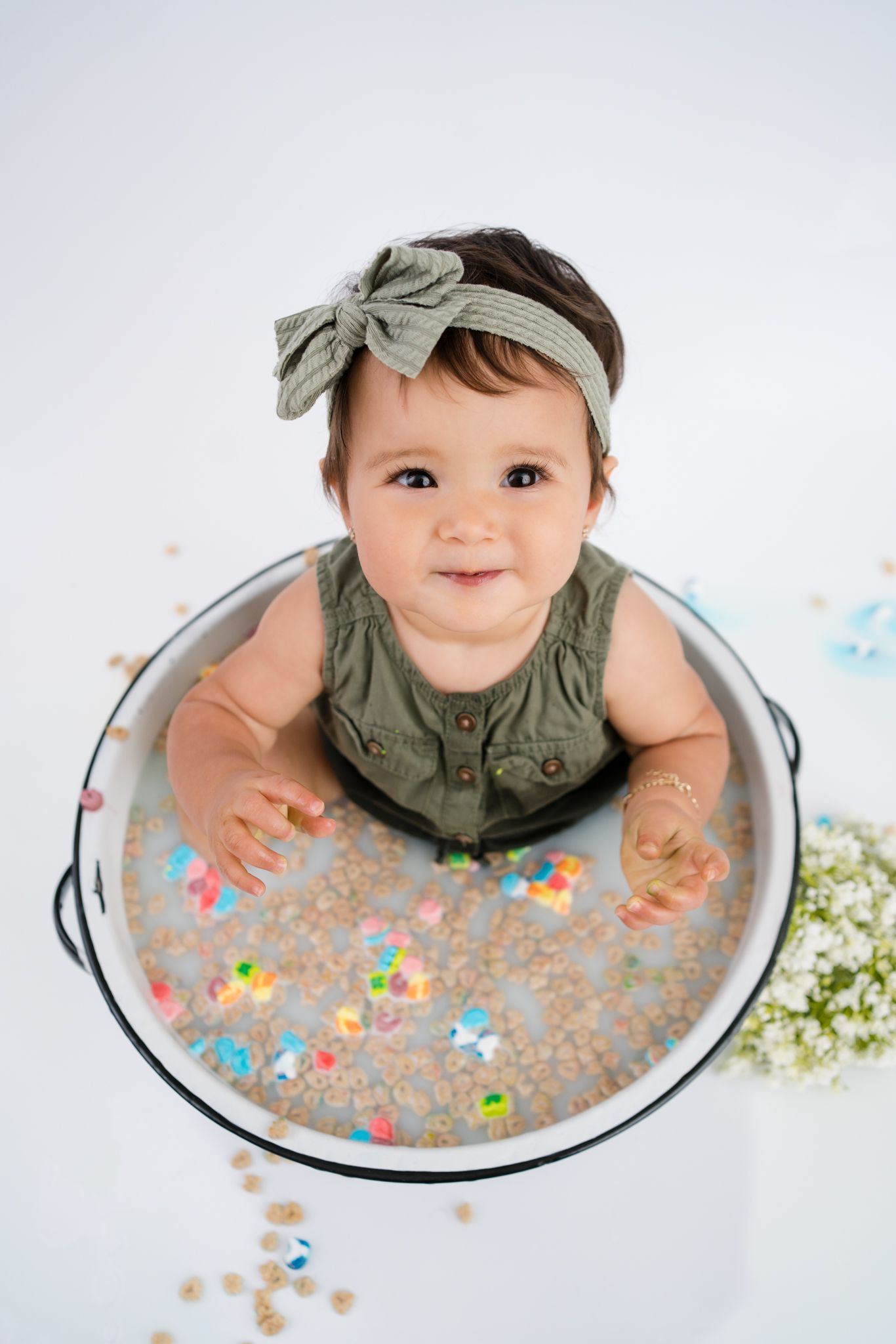 A baby wearing a sage green outfit and headband sits in a metal basin filled with colorful confetti and sparkles.