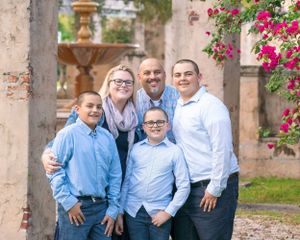 A family of five poses together outdoors near an old stone wall with pink flowers and a fountain in the background.