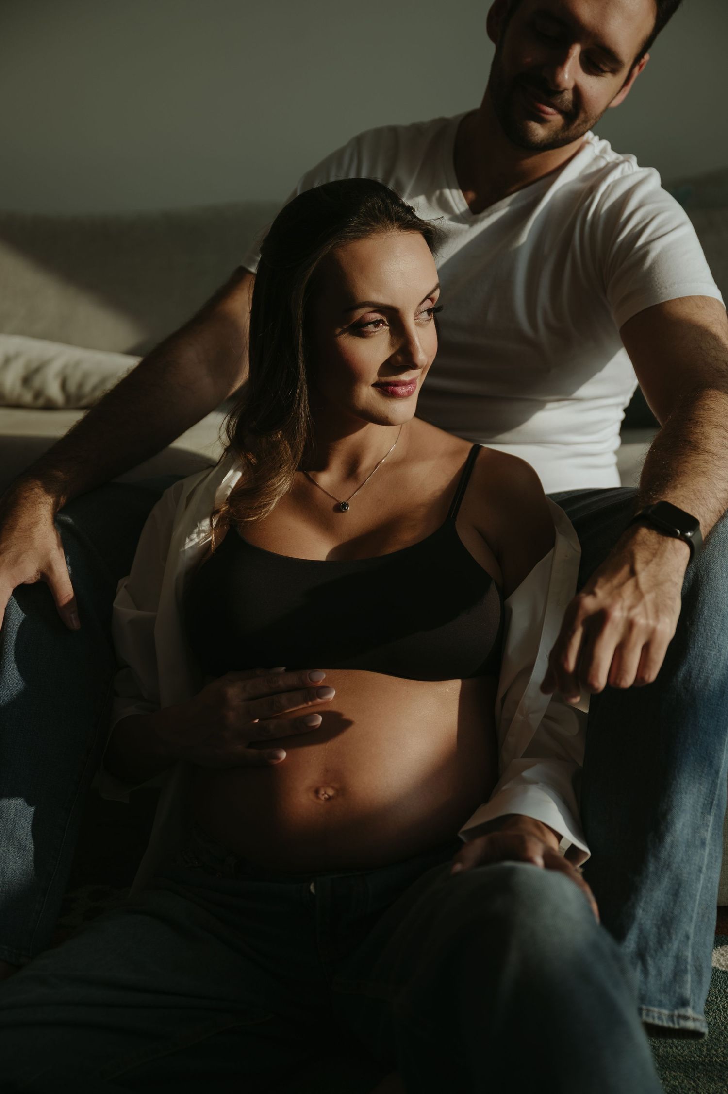 Intimate family moment showing shared laughter during pregnancy photoshoot by window.