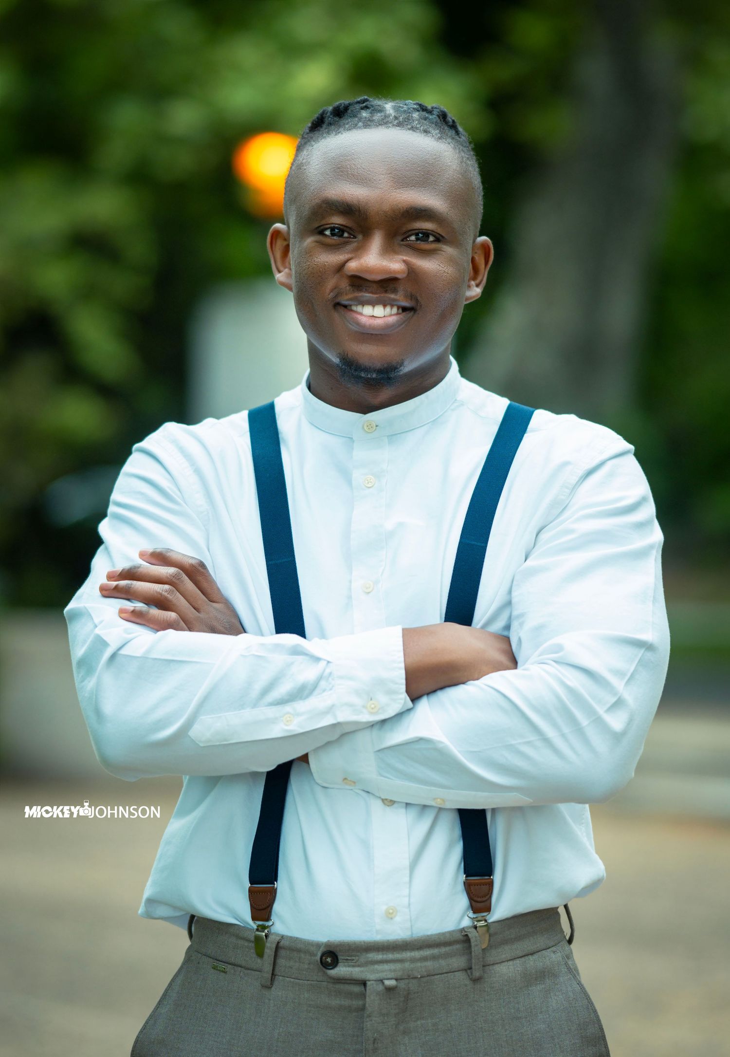 Professional portrait of someone wearing a crisp white dress shirt with navy suspenders against a blurred outdoor background.