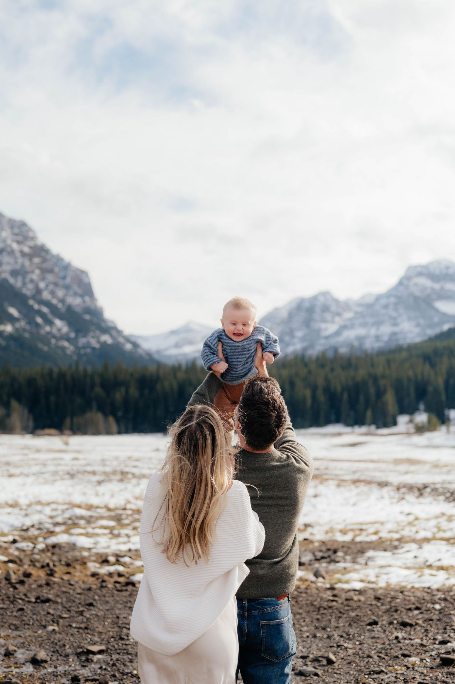 Parents lifting laughing baby in snowy mountain landscape with pine trees and cloudy sky above.