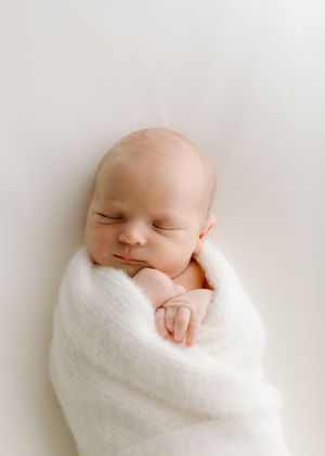 A serene newborn baby swaddled in soft white blanket against neutral background.