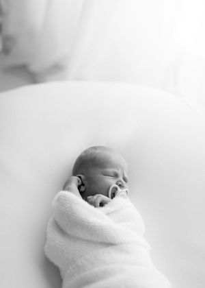 A peaceful newborn wrapped in soft white blanket sleeps soundly against light colored bedding in black and white.