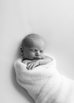 Black and white photo of a peaceful sleeping newborn swaddled in soft white blanket against light background.