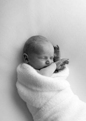 A sleeping newborn baby swaddled in a soft white blanket against a light background in black and white.