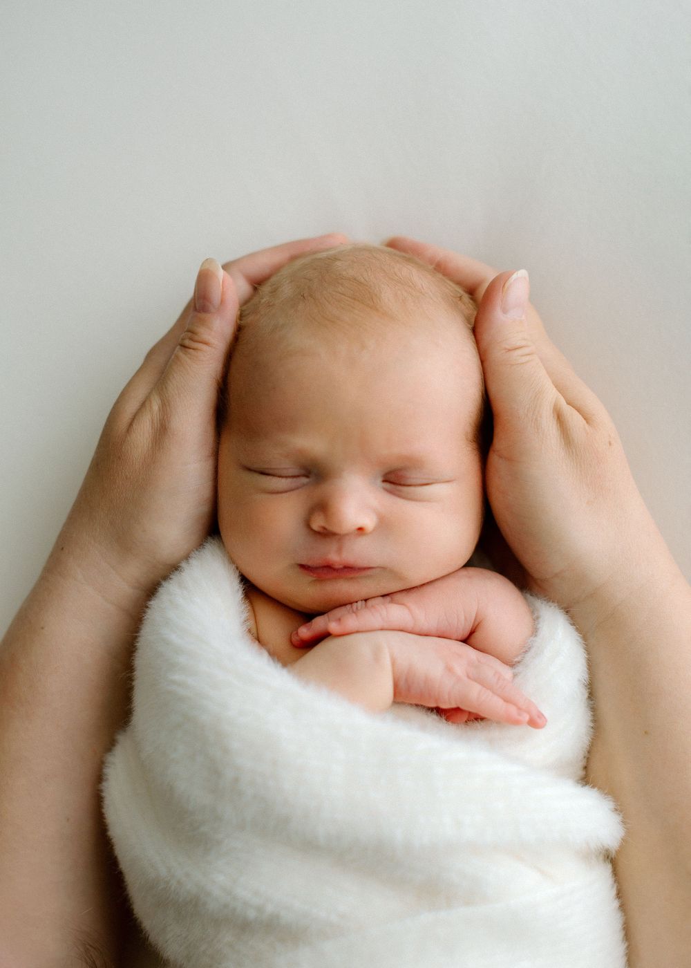 A newborn baby peacefully sleeps while swaddled in a soft white blanket, with gentle hands cradling their head.