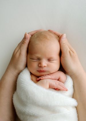 A newborn baby peacefully sleeps while swaddled in a soft white blanket, with gentle hands cradling their head.