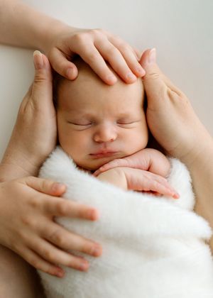 A sleeping newborn wrapped in soft white fabric with gentle hands cradling their head.