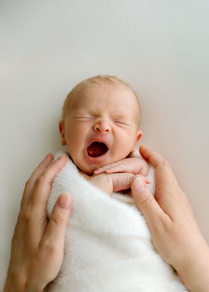 A yawning baby wrapped in white blanket being cradled gently by soft hands on a light background.