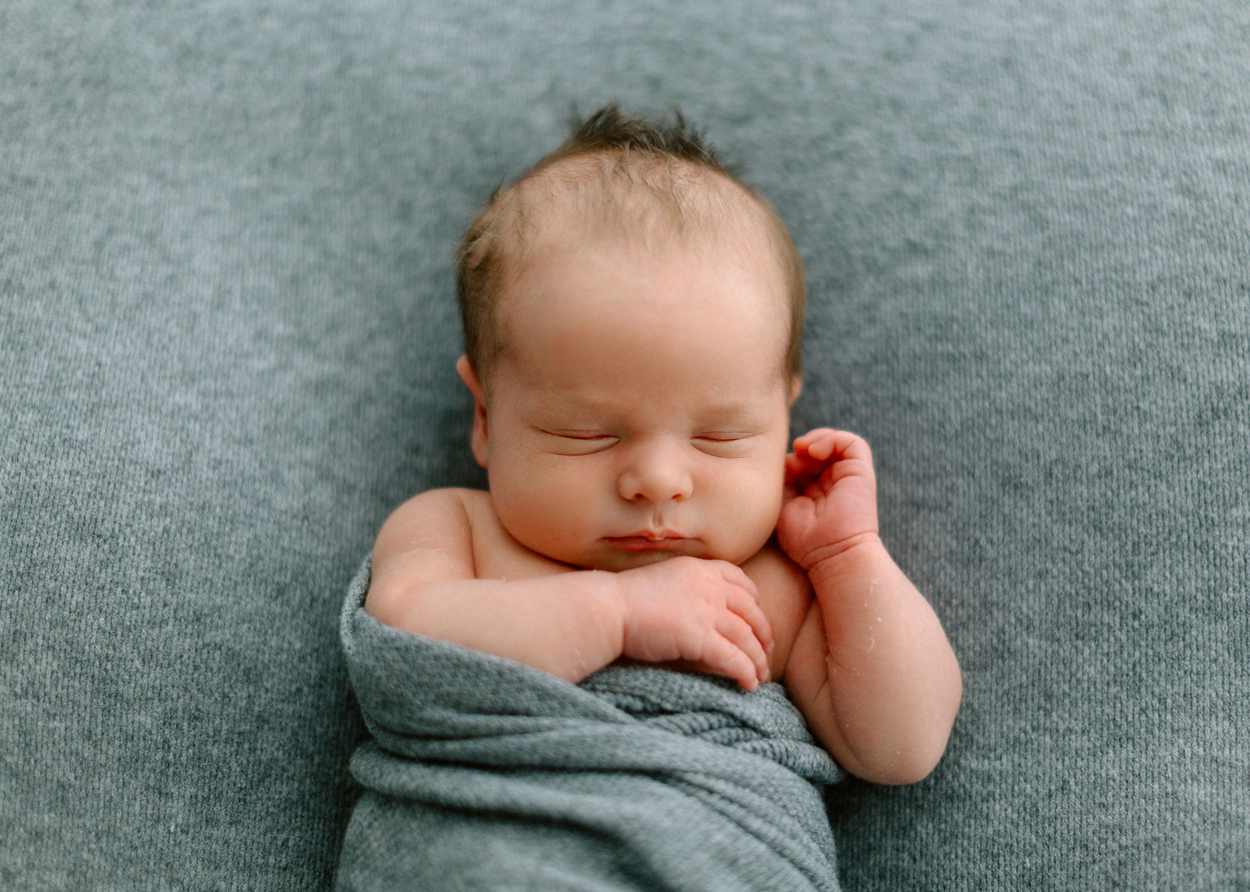 Peaceful sleeping newborn baby wrapped in soft gray blanket on matching gray backdrop.