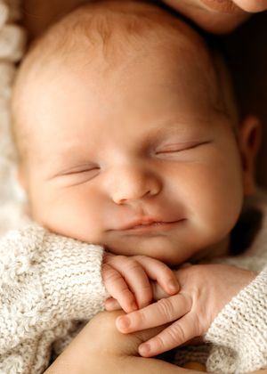 Sleeping newborn baby in knit sweater smiles softly while resting with tiny hands folded.