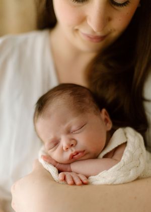 Tender moment of mother holding sleeping newborn baby swaddled in cream colored blanket.