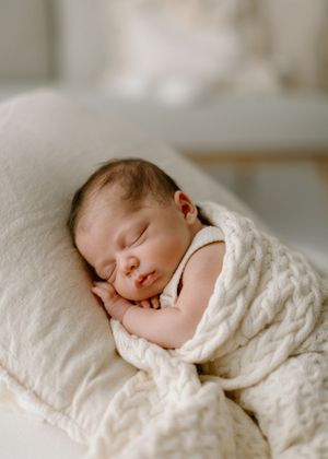 Sleeping newborn baby wrapped in soft white blanket resting peacefully on light colored bedding.