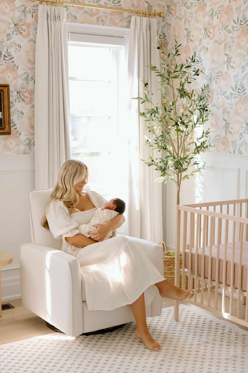 Nursery with floral wallpaper features white glider chair next to window and wooden crib in sunlit corner.