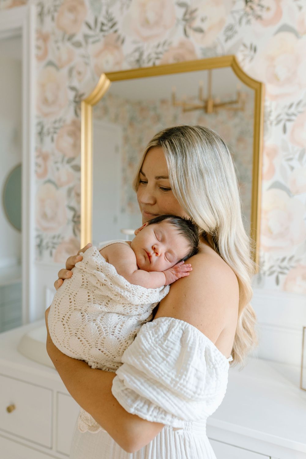 A tender moment captured as a sleeping infant swaddled in white knit rests peacefully against floral wallpaper backdrop.