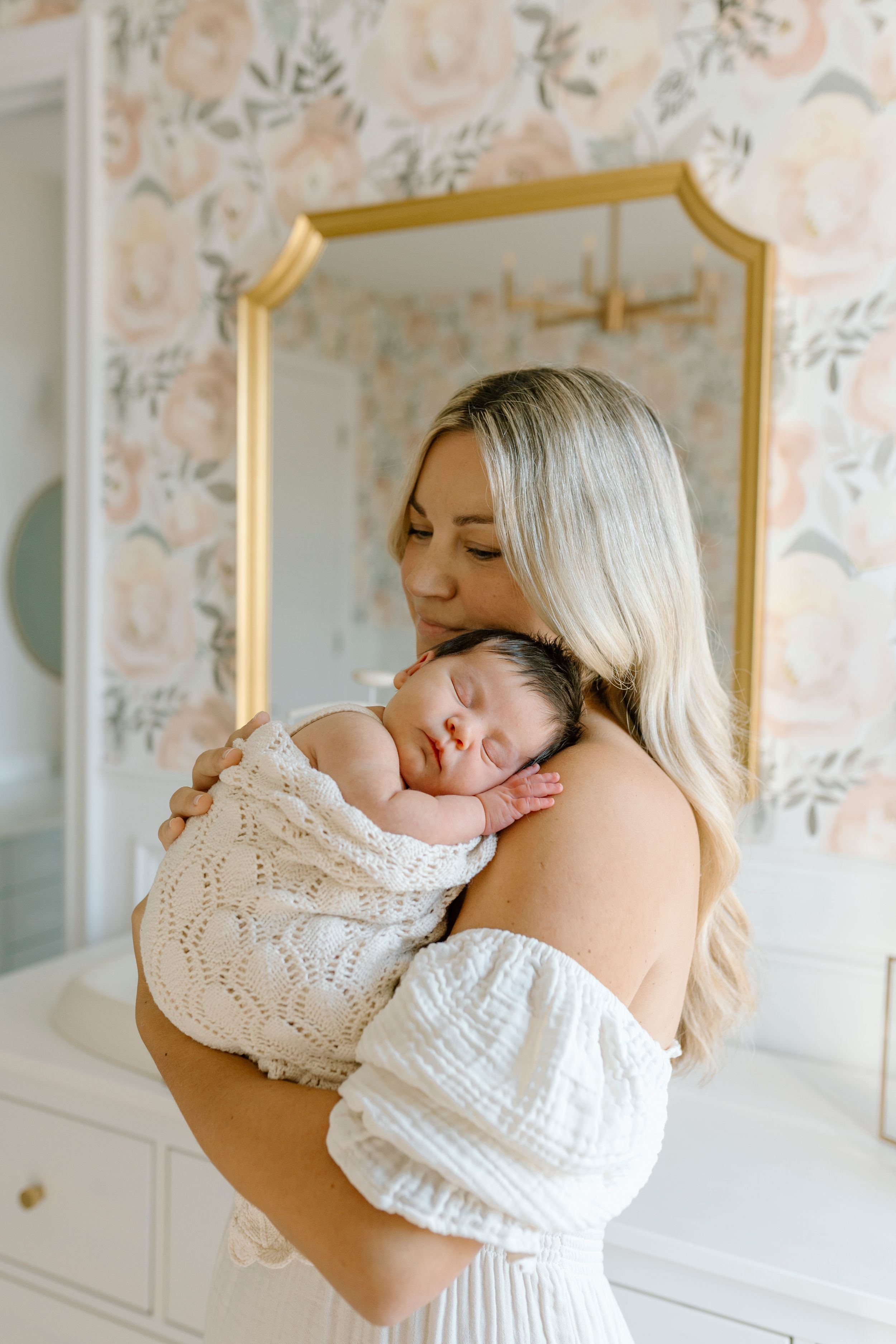 A tender moment captured as a sleeping infant swaddled in white knit rests peacefully against floral wallpaper backdrop.