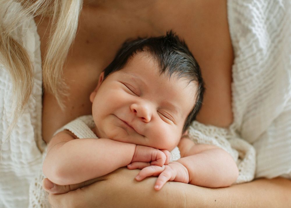 A sleeping newborn baby lies on white bedding while being cradled in gentle hands.