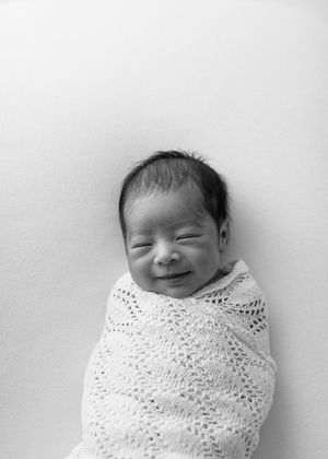 Black and white image of a smiling newborn baby swaddled in a knit blanket against a light background.