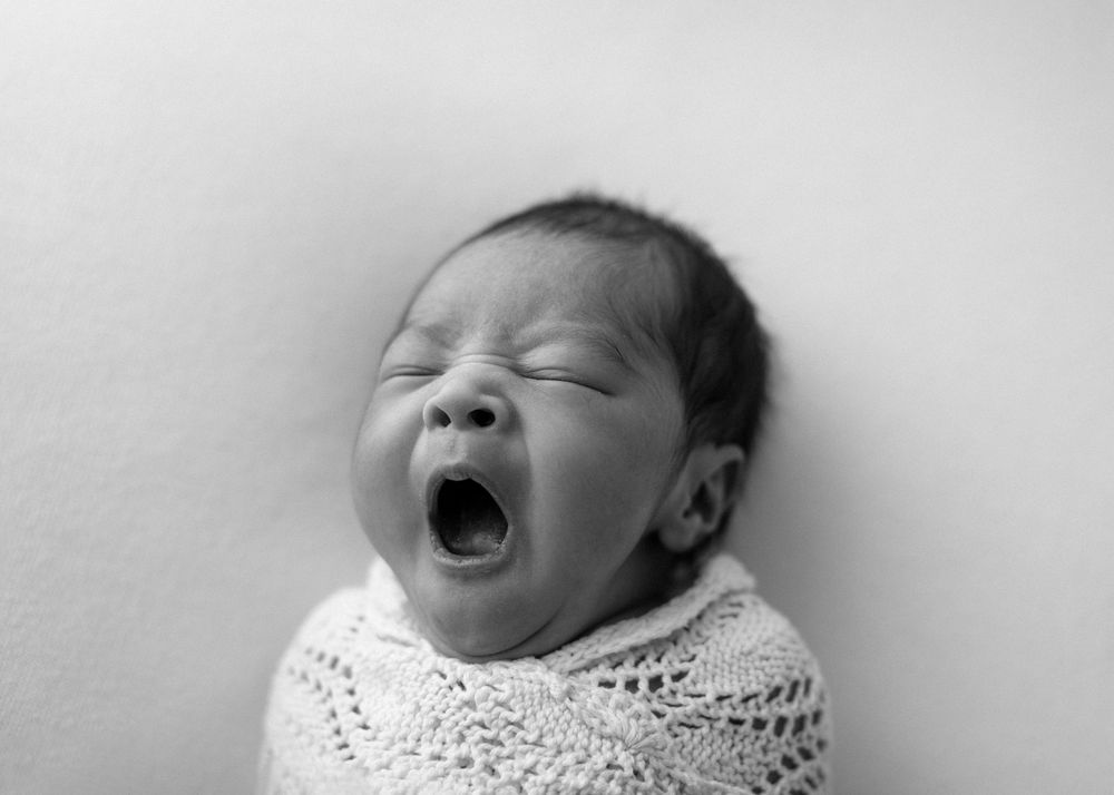 Black and white photo of a newborn baby yawning while swaddled in white lace.