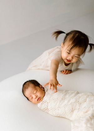Two siblings share a tender moment as one rests in a knitted white blanket while the other smiles with joy.