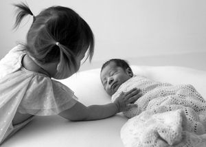 A tender black and white photograph shows a young child reaching out to touch a swaddled newborn baby on a white surface.