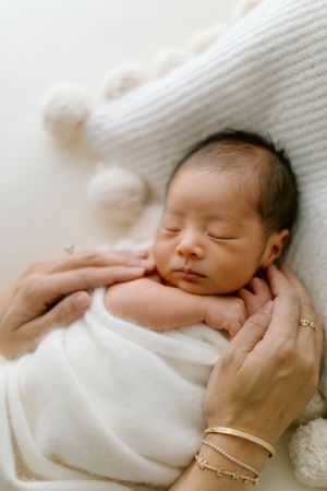 Sleeping newborn wrapped in soft white blanket with gentle hands cradling their delicate face.