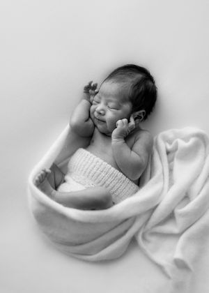 A sleeping newborn baby wrapped in a white blanket and knit wrap lies peacefully in black and white photography.