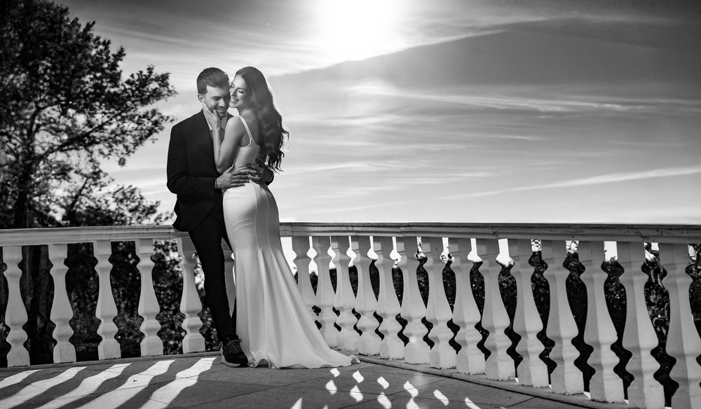 A romantic couple embraces at sunset on a balcony with ornate white stone railings overlooking a scenic vista.