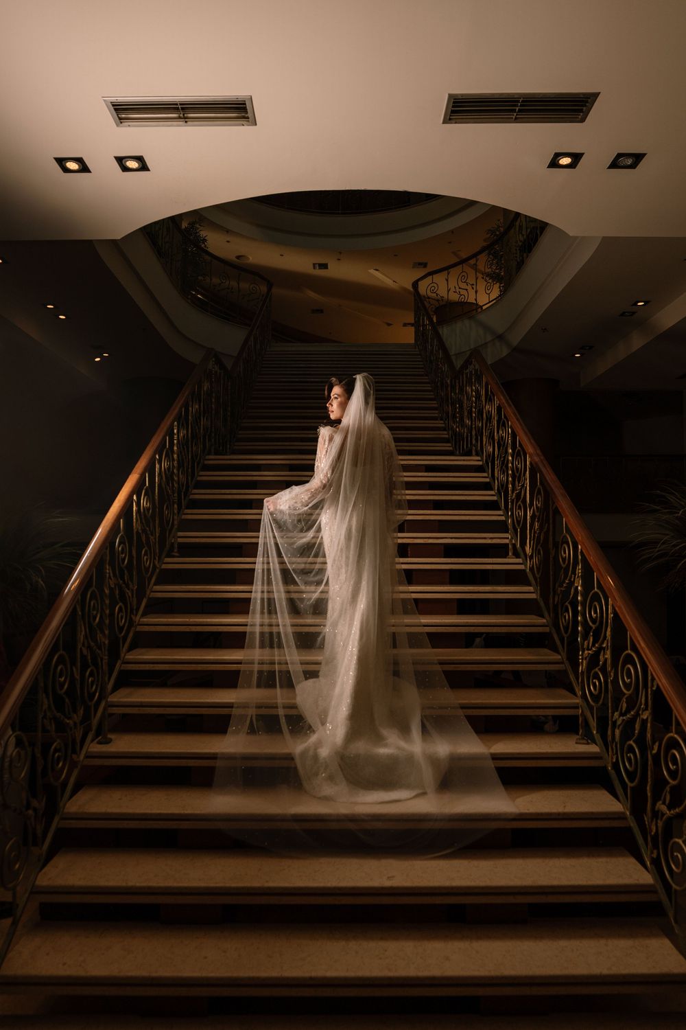 A bride in a flowing white veil poses on an elegant grand staircase in dramatic low lighting creating a moody atmosphere.