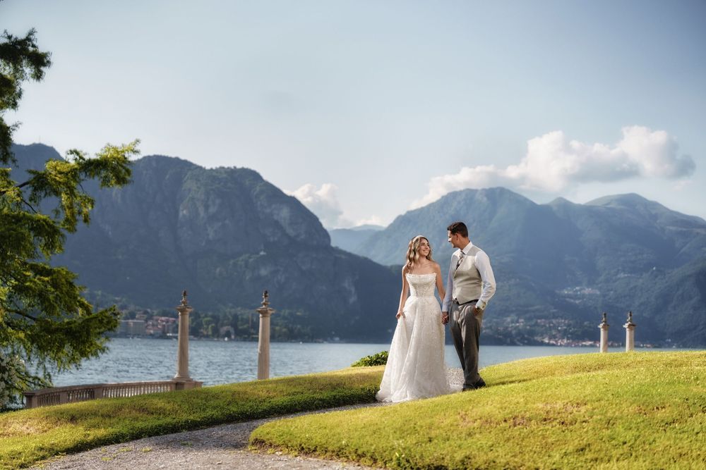 A couple in wedding attire stands on a hilltop overlooking a scenic mountain lake on a beautiful summer day.