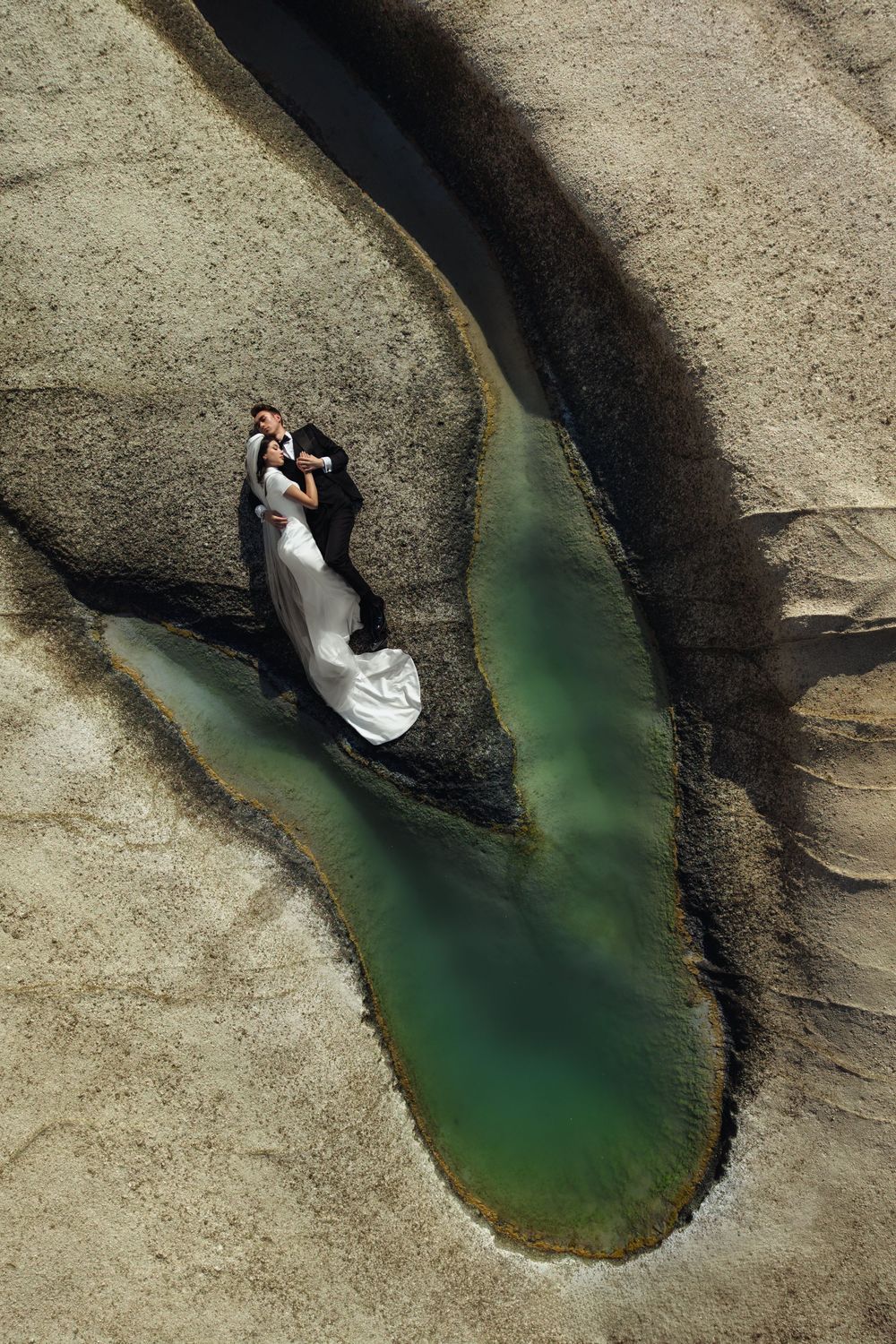 Aerial view of a couple lying on rocky terrain beside a turquoise green pool of water surrounded by stone formations.