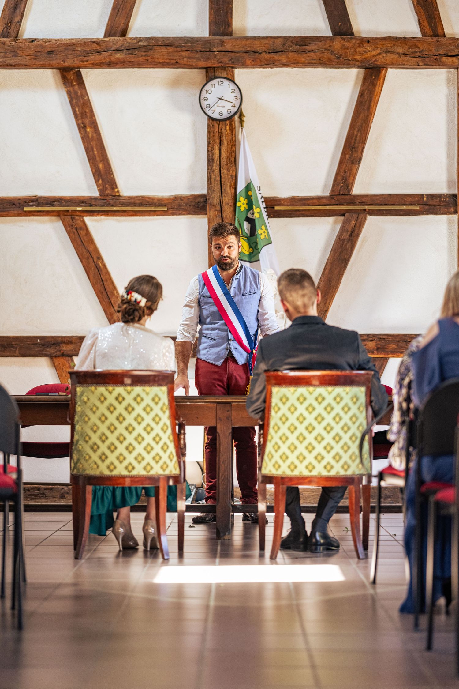 A civil ceremony officiant wearing a tricolor sash stands at the front of a rustic room with timber beams and seated guests.