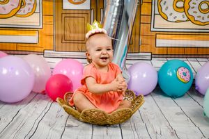 Baby in orange dress and crown sits in basket surrounded by colorful balloons.