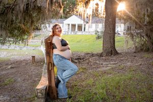 A maternity photo of someone in jeans leaning against a wooden chair outdoors.