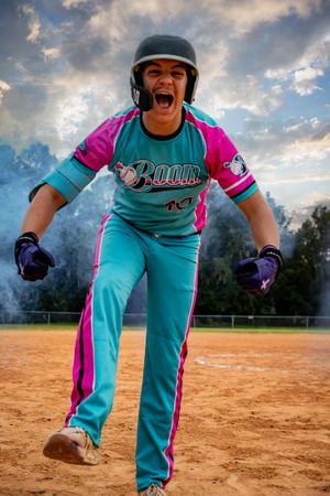 A softball player in a teal and pink uniform celebrates energetically on a dirt field.