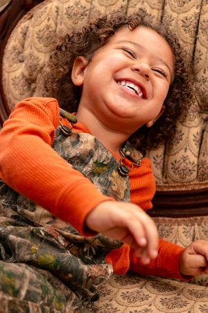 A child in an orange sweater laughs joyfully while laying on a patterned couch.