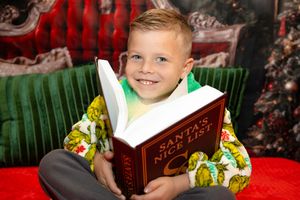 A child sits in a green chair reading a book during the holiday season with Christmas decorations visible in the background.