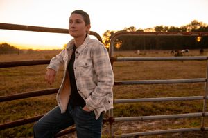 Someone in a plaid jacket leans against a rustic metal fence at sunset in a rural farm setting.
