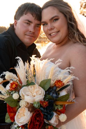 A beautiful autumn wedding bouquet featuring cream roses, red flowers, navy accents and pampas grass in golden sunlight.